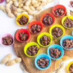 Chocolate-covered peanuts served in colorful paper cups, arranged on a wooden board with raw peanuts scattered around.