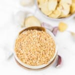 A bowl of dried minced garlic with whole garlic cloves and sliced garlic in the background.