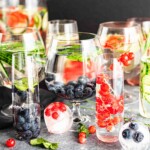 Various glasses filled with fruits like blueberries and red currants, some with added mint leaves, on a gray background.