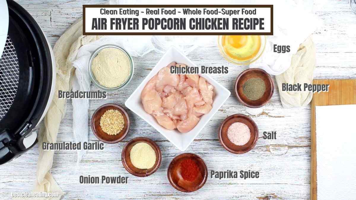 Ingredients for air fryer popcorn chicken recipe are labeled and include chicken breasts, eggs, breadcrumbs, black pepper, salt, paprika spice, onion powder, and granulated garlic, on a wooden surface.