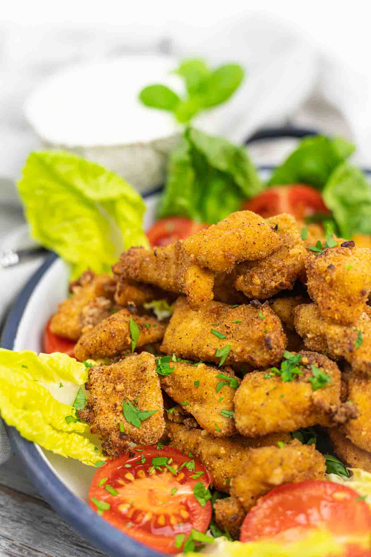 A plate of crispy fried chicken pieces garnished with chopped parsley, surrounded by lettuce leaves and sliced cherry tomatoes, with a small bowl of dipping sauce in the background.