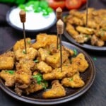 Plates of seasoned, fried chicken bites with garnish and toothpicks for serving. A bowl of dipping sauce and fresh herbs in the background.