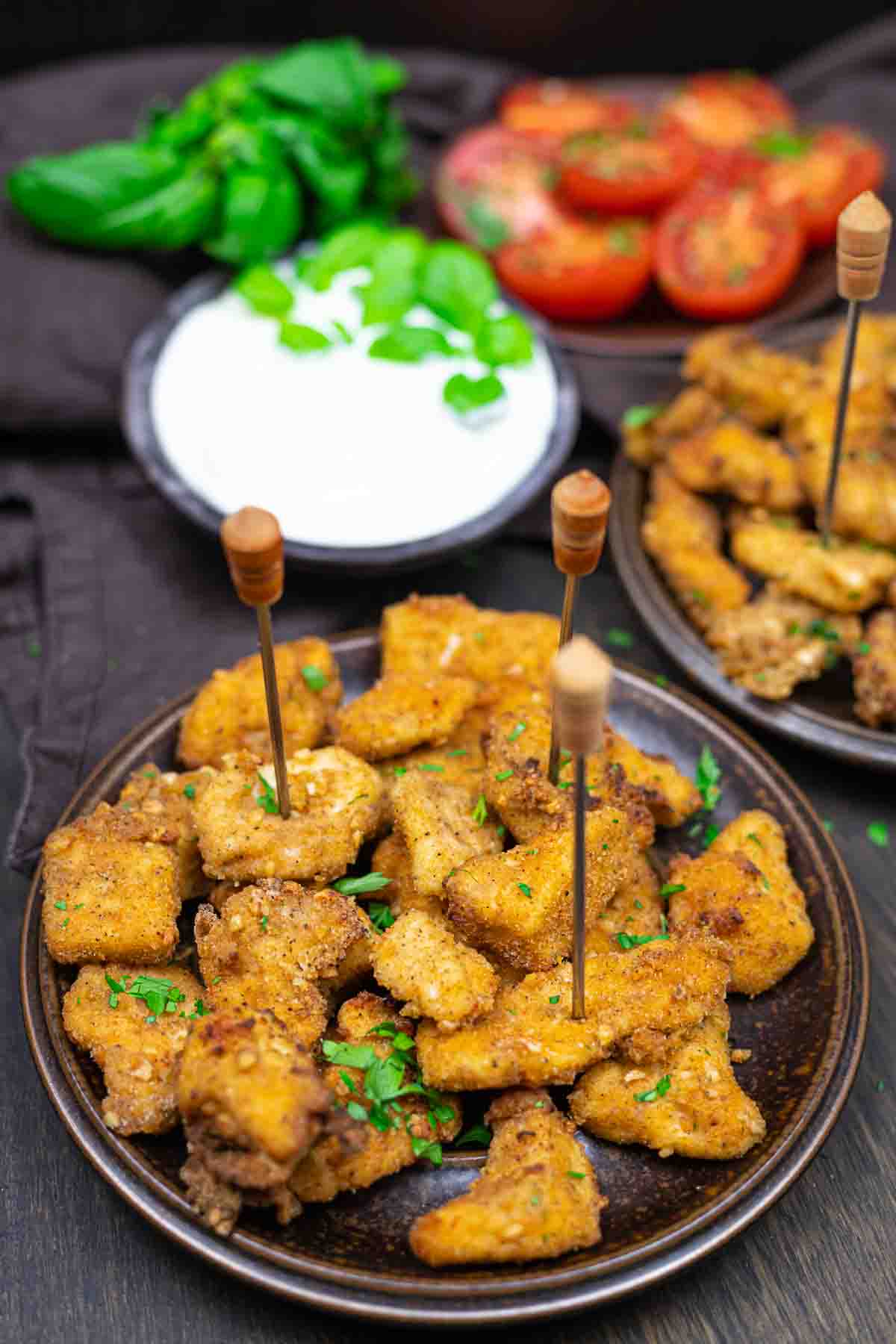 Plates with breaded and fried bites garnished with herbs, accompanied by a dish of sliced tomatoes, basil leaves, and a bowl of white dipping sauce, are arranged on a dark surface. Skewers are inserted into some fish bites.