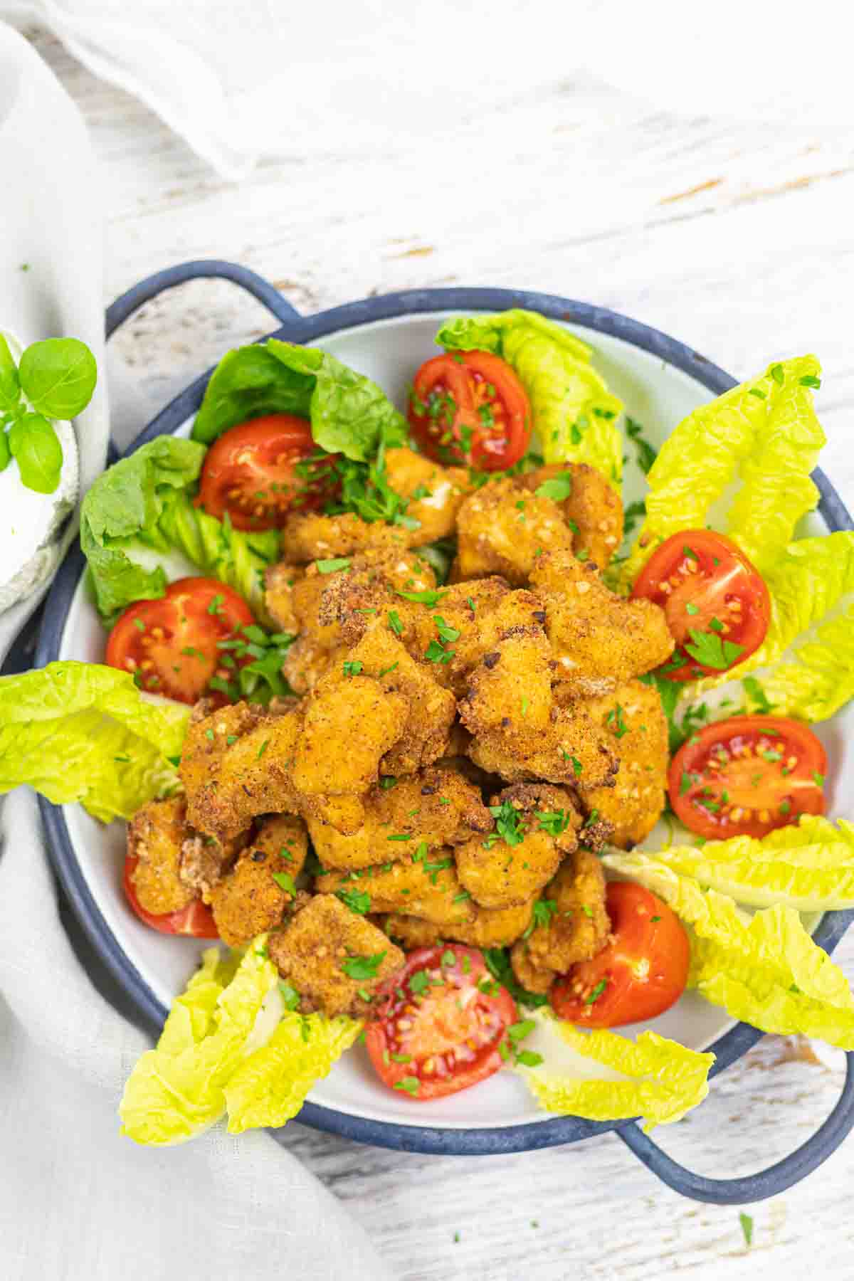 A bowl of crispy fried chicken pieces garnished with chopped parsley, surrounded by fresh cherry tomatoes and lettuce leaves.