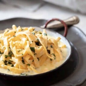 A skillet filled with creamy fettuccine pasta garnished with herbs, placed on a larger plate with a fork resting on the side.