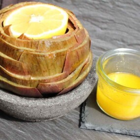 An artichoke with a lemon slice on top is served in a stone bowl next to a small glass jar filled with yellow sauce on a dark slate surface. A fork is placed beside the artichoke.