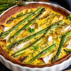 A baked asparagus and cheese quiche in a white dish, garnished with fresh herbs, surrounded by sliced cheese, asparagus spears, and small bowls of seasoning.