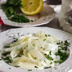 A plate of creamy pasta garnished with fresh parsley, set on a white tablecloth with utensils, a lemon, and red chili peppers in the background.