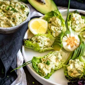 A plate with lettuce leaves filled with a creamy avocado mixture, garnished with sliced green onions, in the center is a halved boiled egg and assorted garnishes, with additional filling in a bowl nearby.
