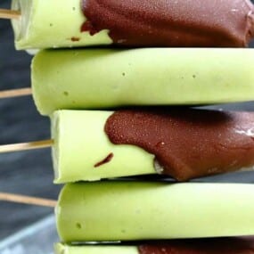 A stack of green popsicles partially dipped in chocolate, each on a wooden stick, arranged vertically.