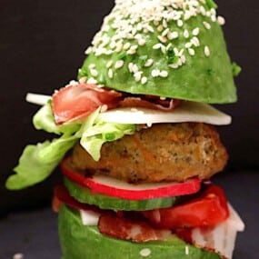 An avocado burger with a patty, lettuce, radishes, tomatoes, and cheese, topped with a sesame-seeded avocado half.