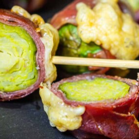 Close-up of brussels sprouts wrapped in prosciutto, held together with a decorative toothpick, with melted cheese on top, arranged on a black surface.