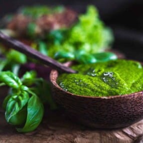 A wooden bowl filled with green pesto sauce, accompanied by fresh basil leaves, placed on a wooden surface.