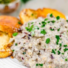 A white plate with creamy sausage dip garnished with chopped herbs, served with slices of toasted bread.