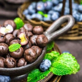 A small bowl of chocolate-covered blueberries garnished with mint leaves sits on a table. In the background, a basket with fresh blueberries and more mint leaves is visible.