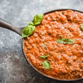 A black skillet filled with tomato-based meat sauce garnished with fresh basil leaves on a textured, gray surface.