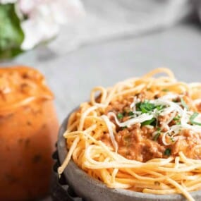 A bowl of spaghetti topped with a meat sauce and garnished with grated cheese and herbs. A jug containing extra sauce is placed next to the bowl.