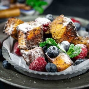 A plate of French toast cubes dusted with powdered sugar, surrounded by fresh raspberries, blueberries, and green mint leaves.