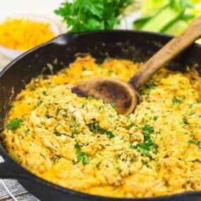 A cast iron skillet filled with a creamy yellow dish garnished with fresh parsley. Wooden spoon inside skillet. Vegetables and shredded cheese visible in the background.