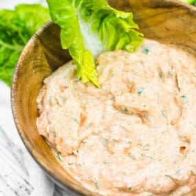 A wooden bowl filled with a creamy dip, garnished with green lettuce leaves, placed on a white wooden surface.