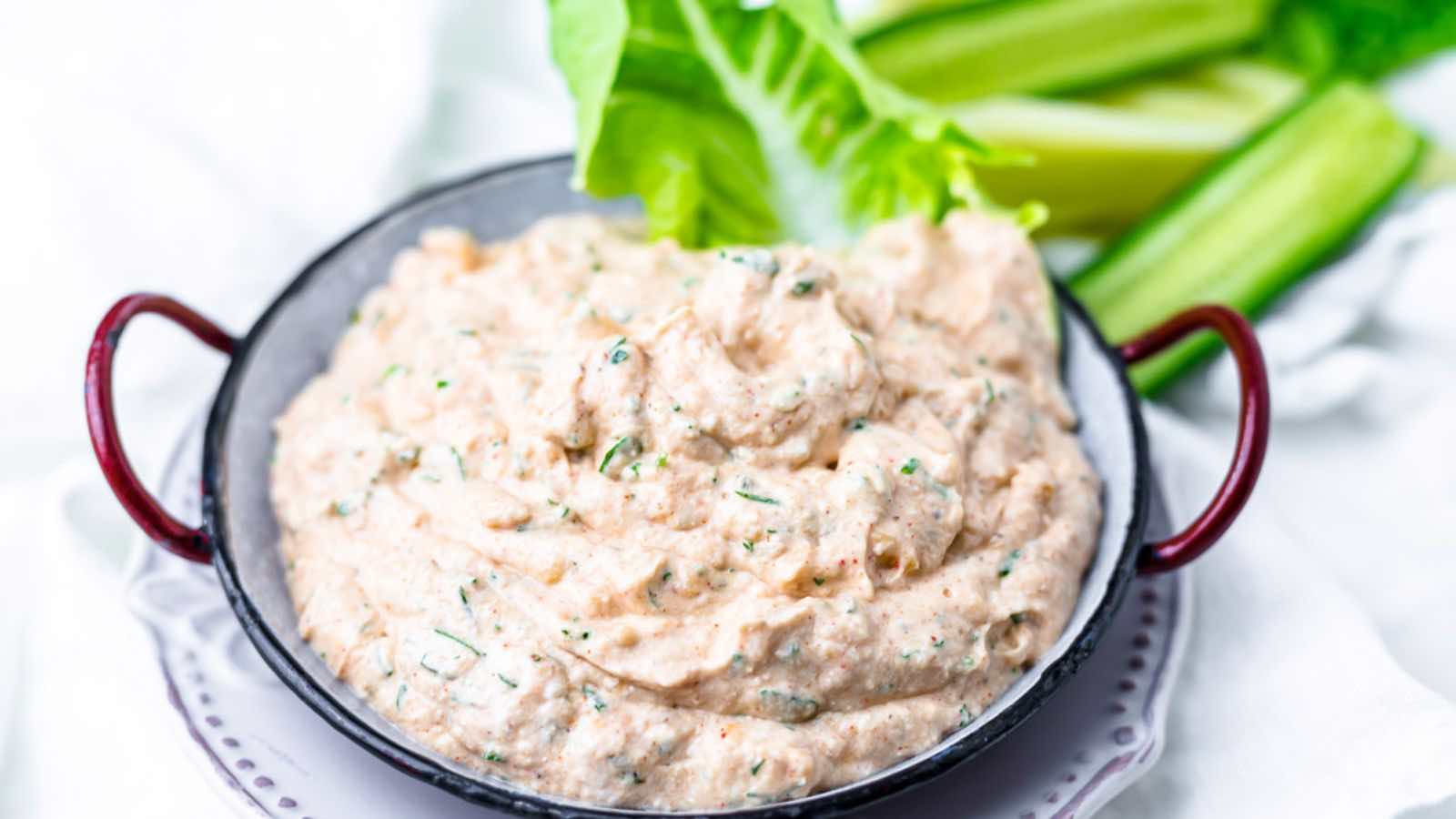 A ceramic bowl with red handles contains a creamy white dip garnished with herbs. Fresh green celery sticks and lettuce leaves are arranged next to the bowl.