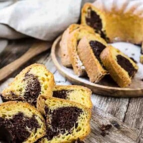 Slices of marble cake arranged on a wooden plate, with a whole marble cake and a linen cloth in the background on a rustic wooden table.
