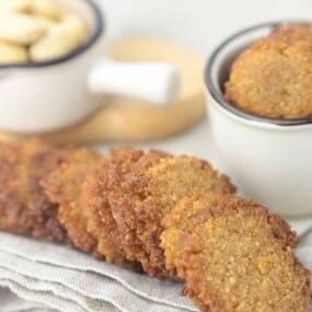 Close-up of a row of crispy brown cookies on a grey cloth, with additional cookies in small white bowls placed in the background.