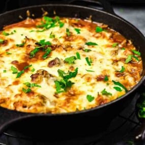 A cast-iron skillet containing a baked dish topped with melted cheese and chopped parsley, with fresh green herbs and shredded cheese nearby.