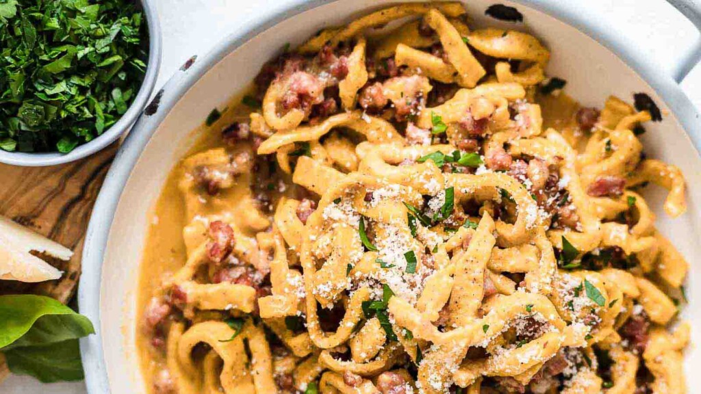 A bowl of creamy pasta with pieces of meat, topped with grated cheese and chopped herbs. A small bowl of chopped herbs is placed beside the pasta.