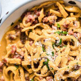 A bowl of creamy pasta with pieces of meat, topped with grated cheese and chopped herbs. A small bowl of chopped herbs is placed beside the pasta.