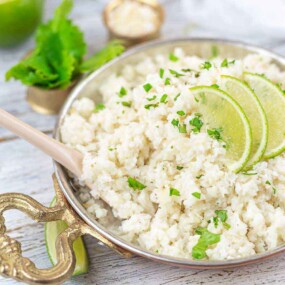 A brass bowl filled with rice garnished with lime slices and cilantro. A wooden spoon rests inside the bowl. A whole lime and a small container with greens are in the background.