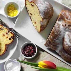 A loaf of cranberry lemon bread, partially sliced, is on a white platter. Beside it are additional slices, a fork, small bowls of lemon zest, dried cranberries, and powdered sugar, next to a tulip.