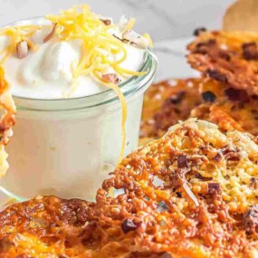A wooden container with crispy cheese crackers spills onto a surface next to a jar of white dip topped with shredded cheese.
