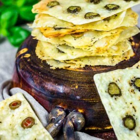 A stack of crispy flatbread with green olives on a wooden board, surrounded by two additional pieces. A sprig of green basil is blurred in the background.