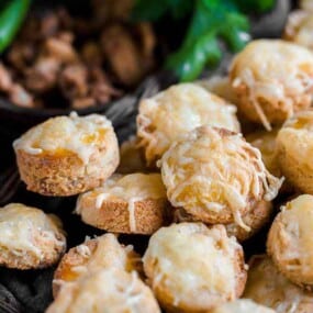 A basket filled with numerous small baked buns topped with melted cheese, with a blurred background of green leaves and nuts.