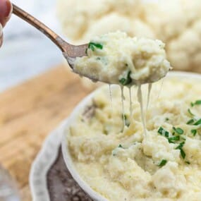 A hand holds a spoonful of creamy, cheesy mashed cauliflower above a bowl filled with the same dish, garnished with chopped parsley. A head of cauliflower is blurred in the background.