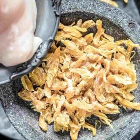 A person is adding raw chicken pieces into a bowl containing dried shrimp.