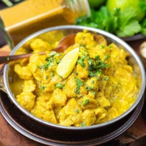 A bowl of yellow curry chicken garnished with a lemon wedge and herbs, placed on a wooden board with a jar of curry sauce, a small bowl of salt, and fresh cilantro in the background.