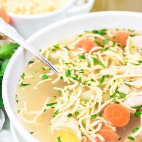 A bowl of chicken noodle soup with chunks of chicken, sliced carrots, parsley, and noodles. A spoon is in the bowl, with fresh parsley and a bowl of shredded cheese nearby.