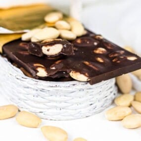 A wicker basket contains a chocolate bar studded with nuts, with additional nuts scattered around the basket.