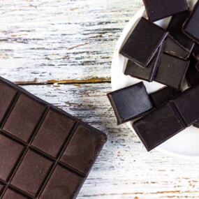 A bar of dark chocolate with square segments is placed on a rustic white wooden surface next to a bowl filled with small dark chocolate pieces.