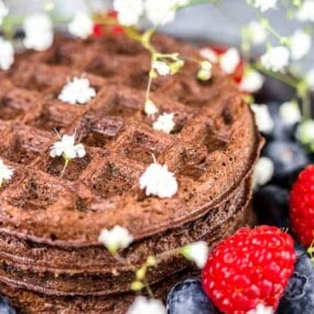 A stack of chocolate waffles on a plate, topped with small white flowers and surrounded by fresh raspberries, blueberries, and green leaves.