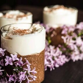 Three glass cups filled with a layered dessert topped with whipped cream and cocoa powder, surrounded by clusters of light purple flowers.