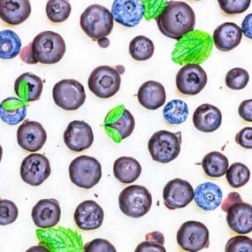 An assortment of chocolate-covered blueberries and fresh blueberries with mint leaves scattered across a light surface.