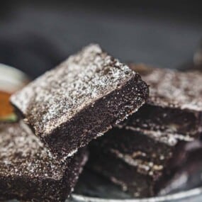 Close-up of a stack of chocolate brownies dusted with powdered sugar, with a bowl of cocoa powder in the background.