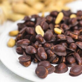 A white plate filled with chocolate-covered peanuts, with a glass bowl of plain peanuts and whole peanuts in the shell in the blurred background.
