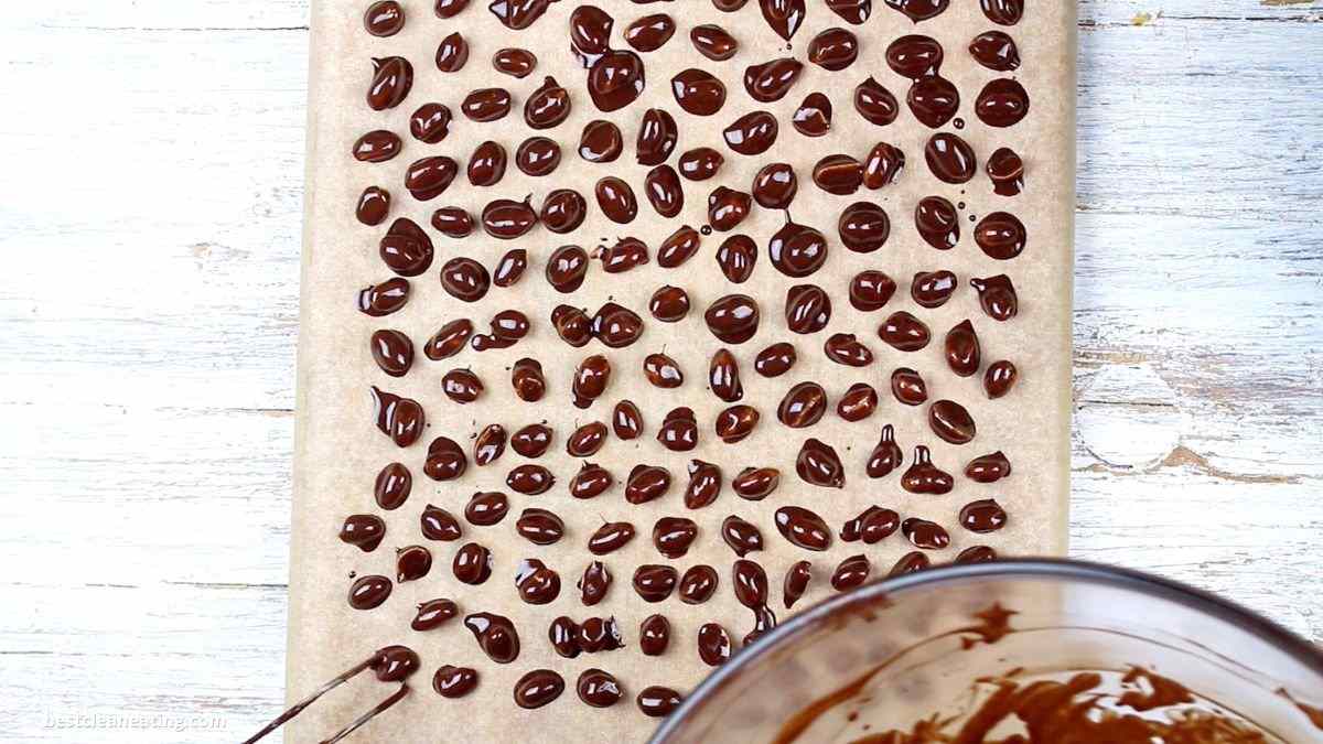 Rows of chocolate-covered almonds spread out on parchment paper, with a bowl of melted chocolate visible in the corner, set on a rustic wooden background.