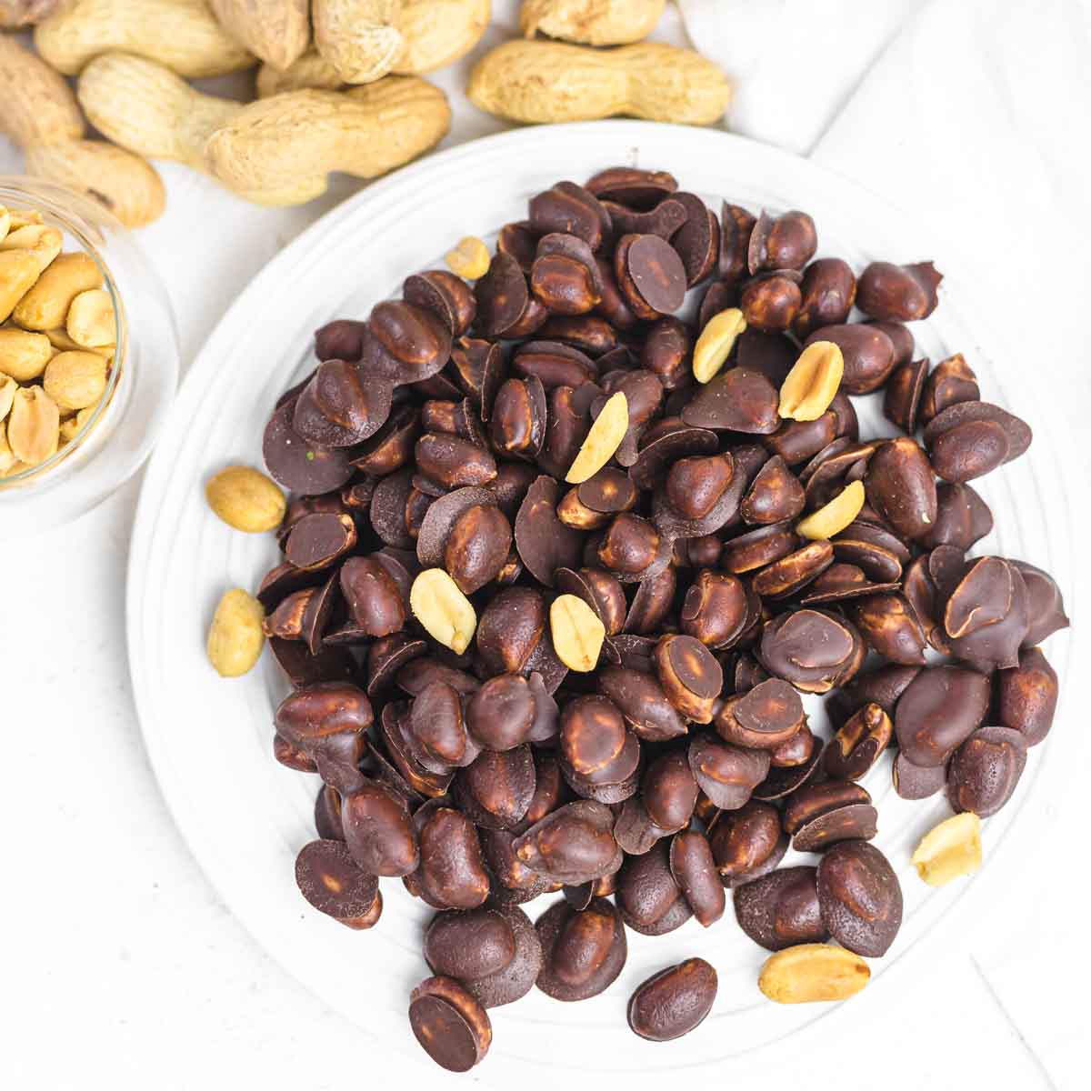 A plate of salted peanuts coated in dark chocolate, with a few uncoated peanuts scattered around, on a white surface.