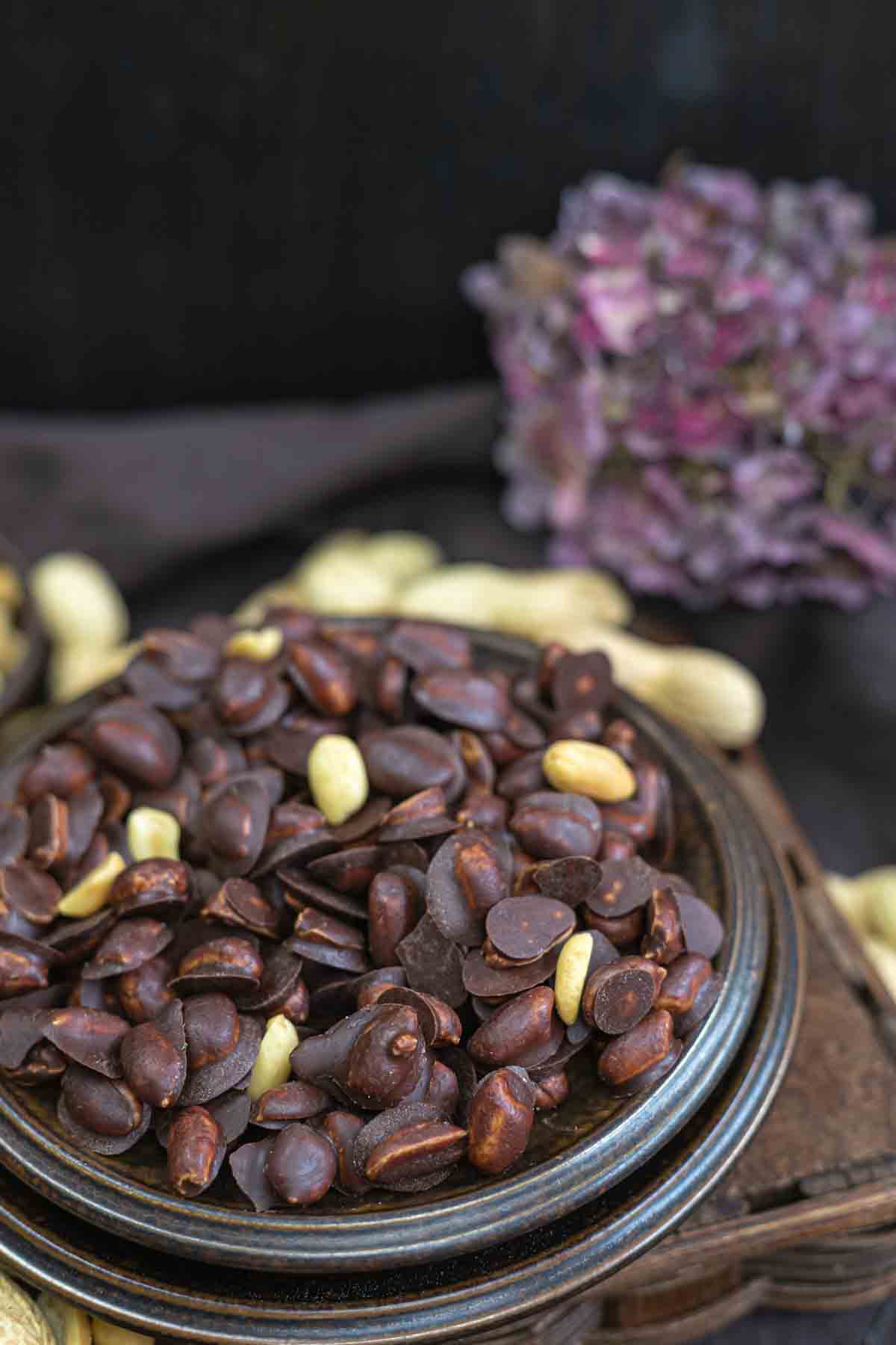 A bowl of chocolate peanuts on a dark wooden surface, with a purple hydrangea flower in the background.