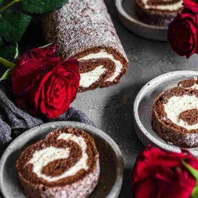 Chocolate Swiss rolls with a cream filling are arranged on plates, accompanied by vibrant red roses on a grey surface.